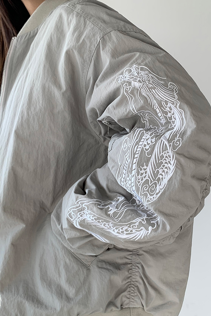 White bomber jacket with beige and grey tones featuring intricate white dragon embroidery, by TM3, displayed on a hanger.